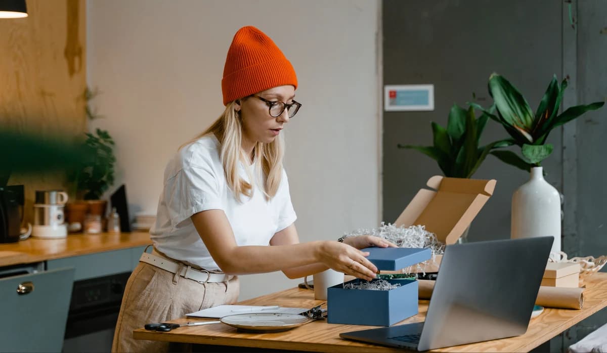 A shop owner packing an order at a desk beside a laptop